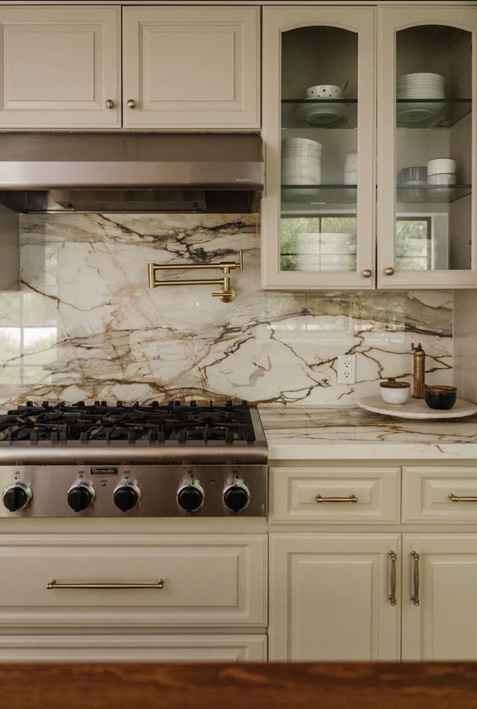 Marble slab backsplash with brass pot filler above a professional range, cream cabinetry